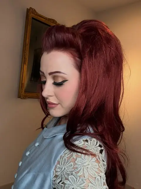 Marguerite Anderson in profile, auburn hair pinned up in a 1960s style, wearing a powder-blue dress with lace sleeves, a gilt-framed painting behind her